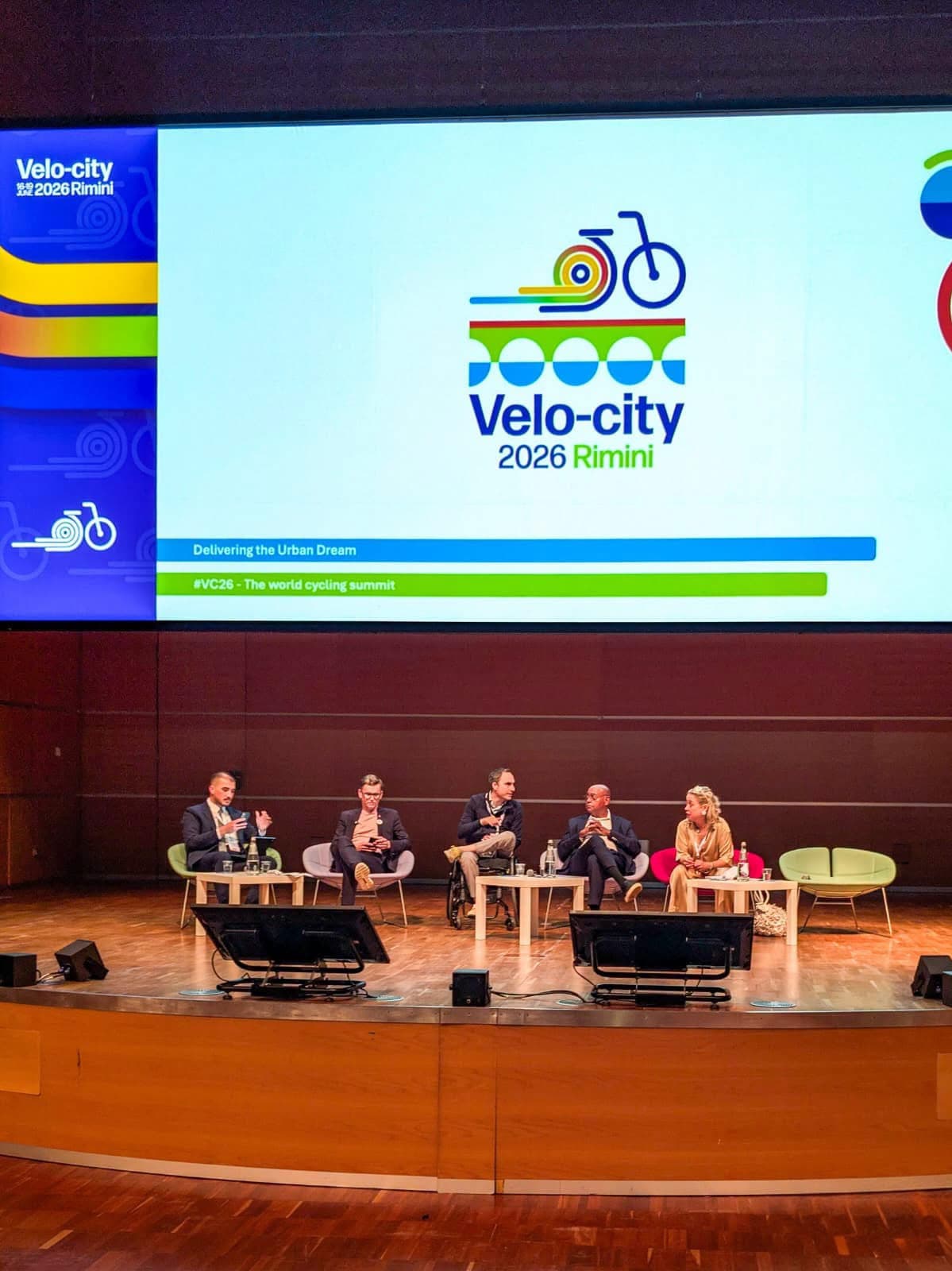 Panel di Velo-city 2026 al Palacongressi di Rimini: cinque relatori seduti sul palco davanti a uno schermo con il logo dell'evento e il tema 'Delivering the Urban Dream'.